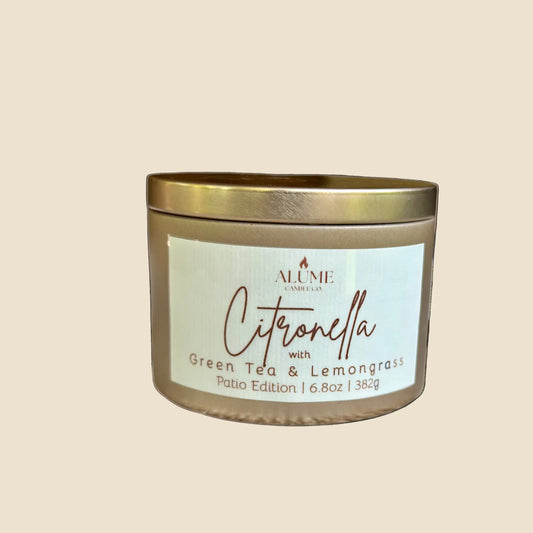 A gold candle with a label that reads 'Citronella with Green Tea & Lemongrass', set on a concrete surface with a background that includes dried plants and a lit candle.