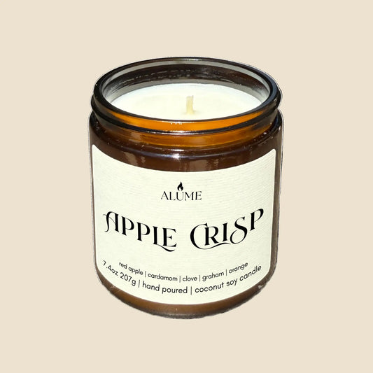 Candle in a glass jar labeled 'Apple Crisp' by Alume on a white background