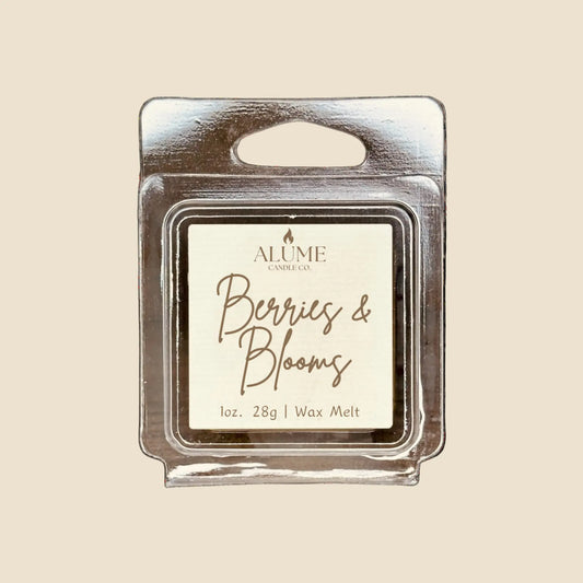 A wax melt pack labeled 'Berries & Blooms' from Alume, with a weight of 1 oz (28g). The pack is transparent, revealing a white wax inside, and it's set against a background with a blurred out candle.
